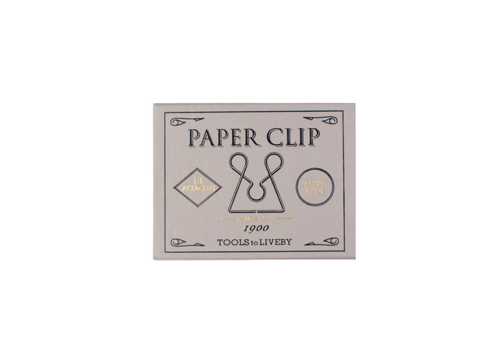 A top-down view of the closed, vintage-inspired light grey sliding matchbox for the TOOLS to LIVEBY McGill paper clip, showcasing elegant black typography with gold foil-stamped accents detailing its 1900 heritage design.