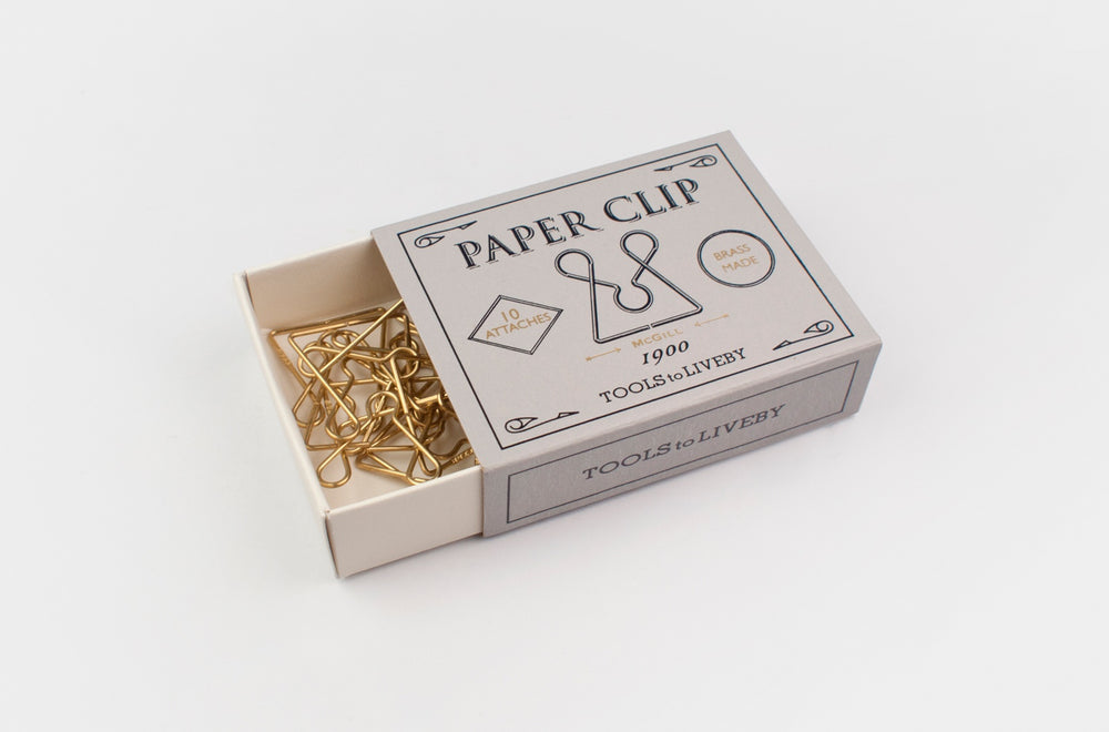 An open vintage grey sliding matchbox for the TOOLS to LIVEBY McGill paper clip, revealing a tangled collection of gleaming raw brass clips inside, resting on a white background.