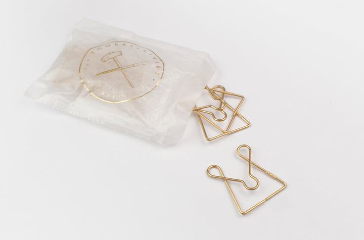 A semi-transparent wax paper bag with a gold foil-stamped TOOLS to LIVEBY logo, from which several McGill brass paper clips are spilling onto a white surface.