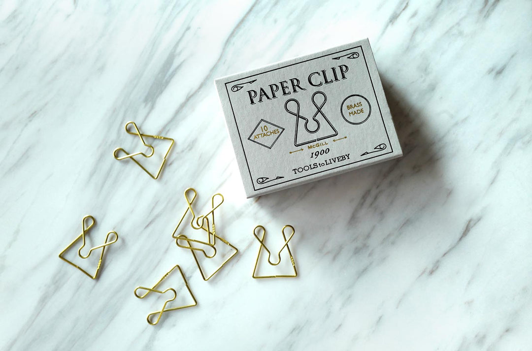 A lifestyle shot featuring a closed TOOLS to LIVEBY McGill brass paper clip box positioned on a bright white marble surface, surrounded by several scattered brass paper clips that highlight their unique wirework pattern.