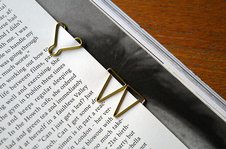 A detailed close-up photograph of a McGill brass paper clip securely holding together pages of an open book, contrasting the gold metal against the white paper with text.