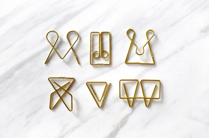 A top-down view of six unique TOOLS to LIVEBY brass paper clips laid out side-by-side on a white marble surface, including the McGill, Mogul, Owl, Niagara, Ideal, and Weis designs, with the subtle "TTLB" branding visible on the wire.