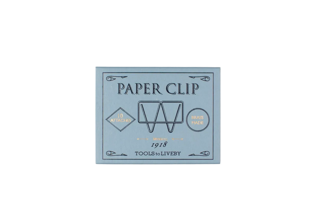 A direct top-down product shot of the closed, slate-blue sliding matchbox for the TOOLS to LIVEBY Mogul brass paper clip, showcasing the elegant black typography and gold foil-stamped accents detailing its 1918 heritage.