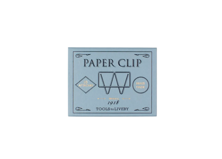 A direct top-down product shot of the closed, slate-blue sliding matchbox for the TOOLS to LIVEBY Mogul brass paper clip, showcasing the elegant black typography and gold foil-stamped accents detailing its 1918 heritage.