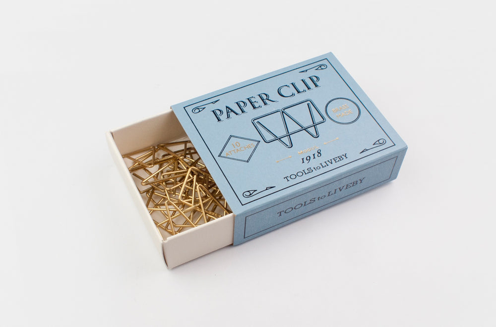 An open vintage-style slate-blue sliding matchbox revealing a tangled cluster of gleaming raw brass Mogul paper clips resting in the tray against a clean white background.