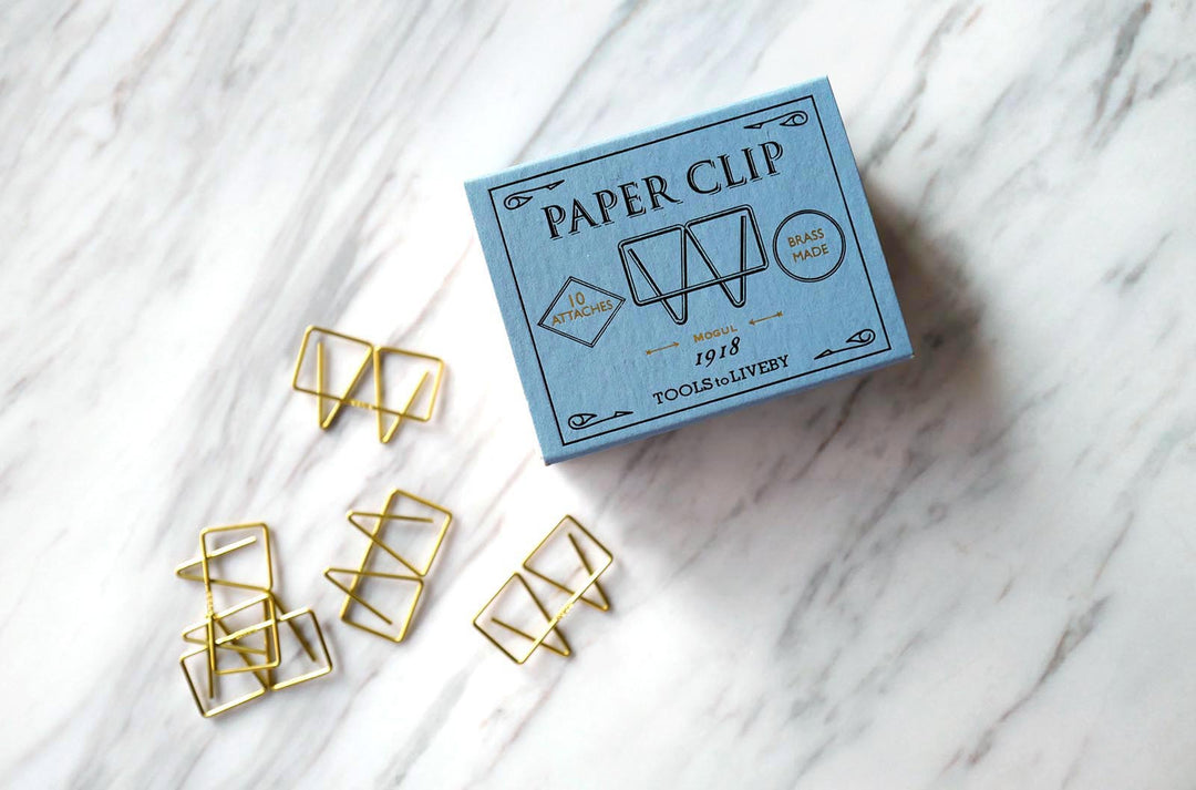 A lifestyle shot on a white marble surface featuring the closed, slate-blue sliding matchbox packaging for the Mogul brass paper clip, surrounded by five loose clips arranged to highlight their geometric wire form.
