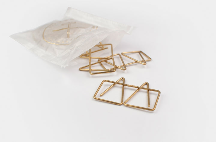 Top-down view of several TOOLS to LIVEBY Mogul brass paper clips spilling elegantly onto a white surface from a semi-transparent wax paper bag featuring a gold foil-stamped TTLB logo.