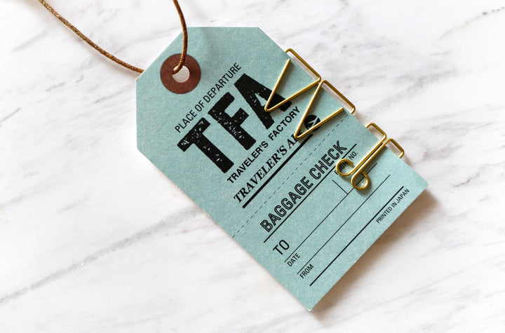 A close-up lifestyle photograph of a blue-green Traveler's Notebook baggage check tag secured to a document with three different unique brass paper clips, including the Mogul design, resting on a white marble surface.