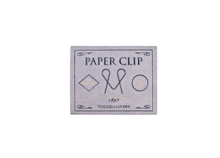 A direct top-down product shot of the closed, lavender sliding matchbox for the TOOLS to LIVEBY Niagara brass paper clip, showcasing elegant black typography and gold foil-stamped accents detailing its 1897 heritage.