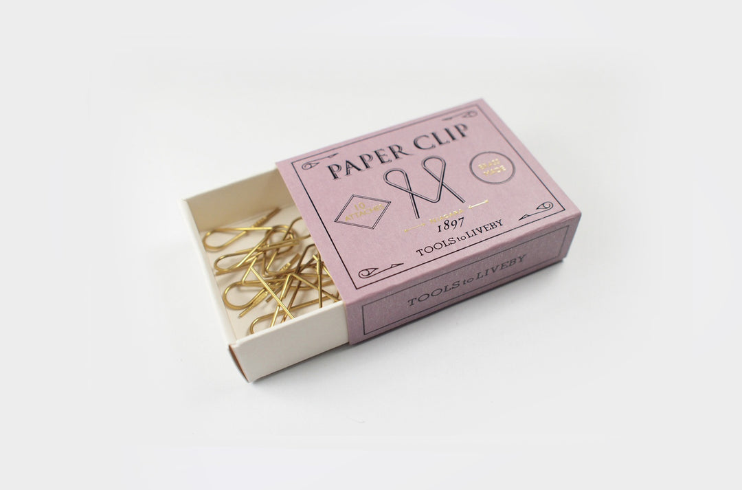 An open vintage-style lavender sliding matchbox revealing a tangled cluster of gleaming raw brass Niagara paper clips resting in the tray against a clean white background.