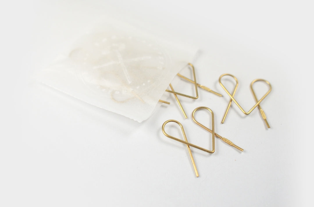 Top-down view of several heart-shaped TOOLS to LIVEBY Niagara brass paper clips spilling elegantly onto a white surface from a semi-transparent wax paper bag featuring a subtle TTLB logo.