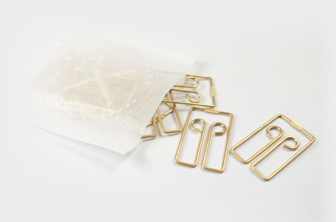 Top-down view of several TOOLS to LIVEBY Owl brass paper clips spilling elegantly onto a white surface from a semi-transparent wax paper bag featuring a gold foil-stamped TTLB logo.
