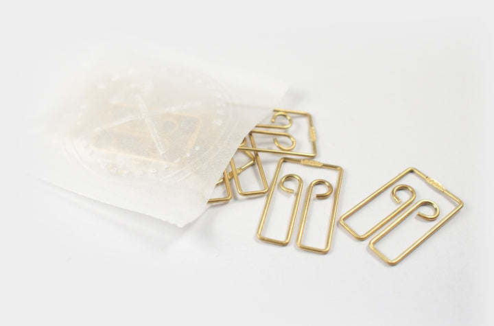 Top-down view of several TOOLS to LIVEBY Owl brass paper clips spilling elegantly onto a white surface from a semi-transparent wax paper bag featuring a gold foil-stamped TTLB logo.