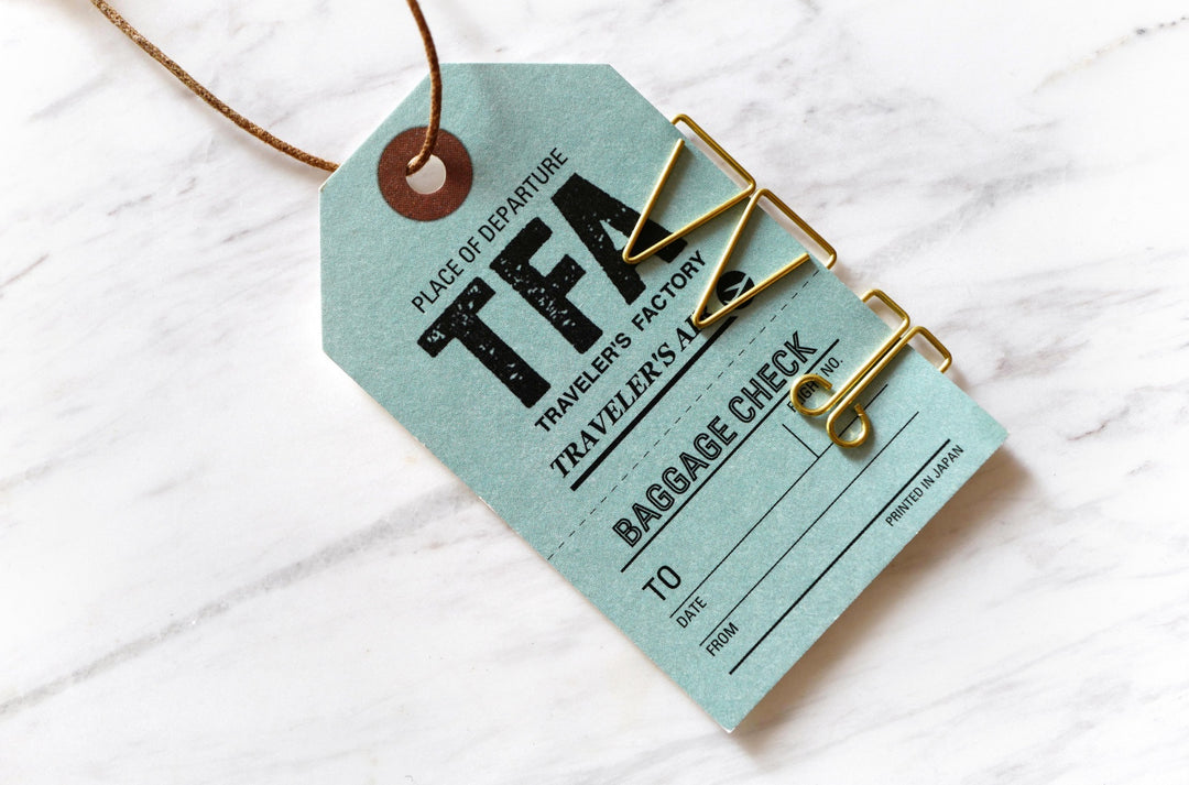 A close-up lifestyle photograph of a blue-green Traveler's Notebook baggage check tag secured to a document with three different unique brass paper clips, resting on a white marble surface.
