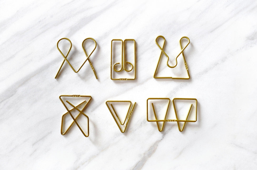 A clean, symmetrical grid layout on a white marble background displaying six unique TOOLS to LIVEBY brass paper clips side-by-side, including the Mogul, McGill, Owl, Niagara, Ideal, and Weis designs.