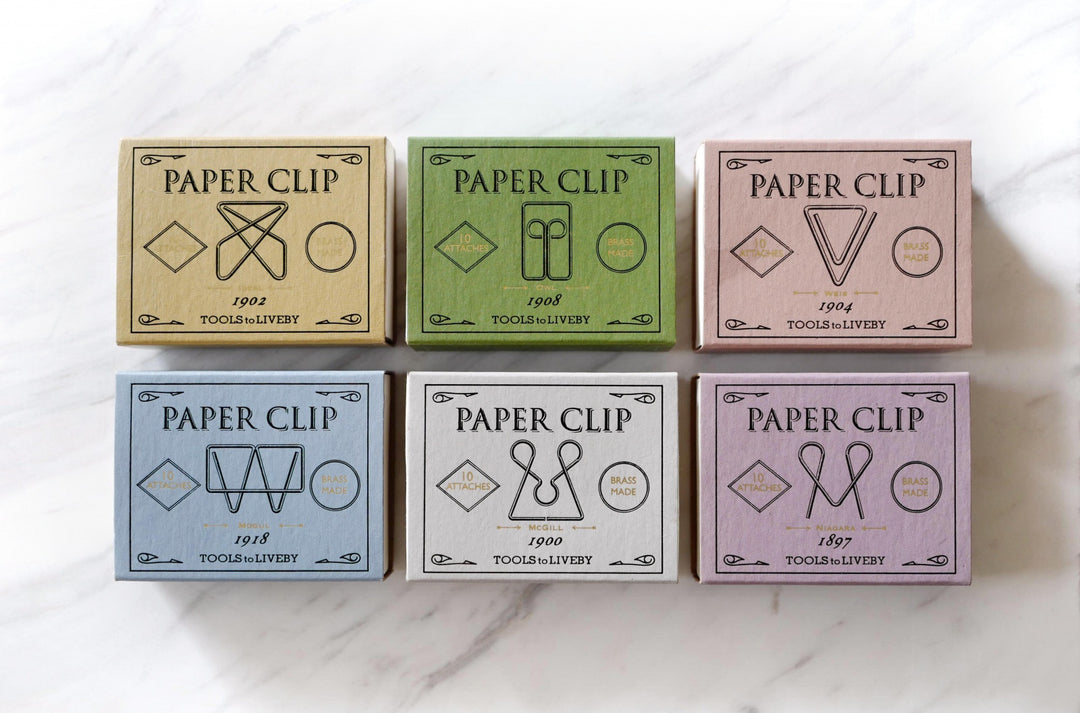 A top-down grid view of six unique TOOLS to LIVEBY brass paper clip boxes arranged neatly on a white marble surface, showcasing their vintage matchbox design in distinct muted colors including the emerald green Owl box.