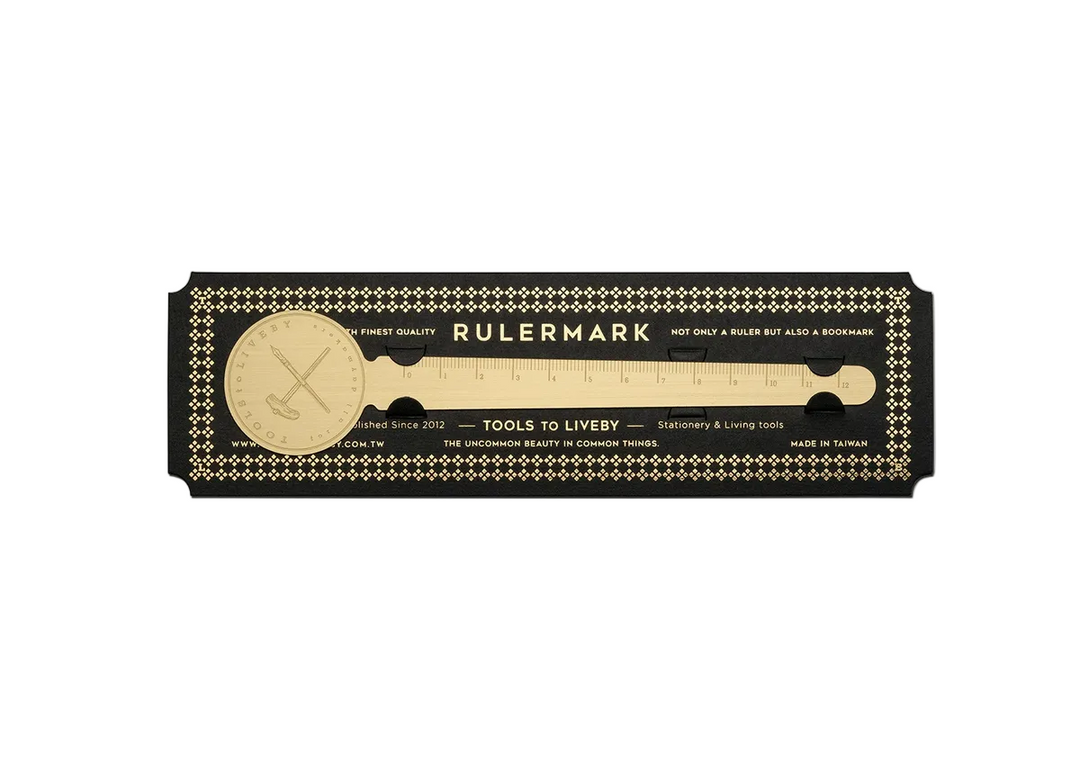 A clean, direct top-down product shot of the TOOLS to LIVEBY Brass Rulermark securely held within its premium black cardstock carrier, featuring vintage gold foil typography and a repeating diamond border against a stark white background.