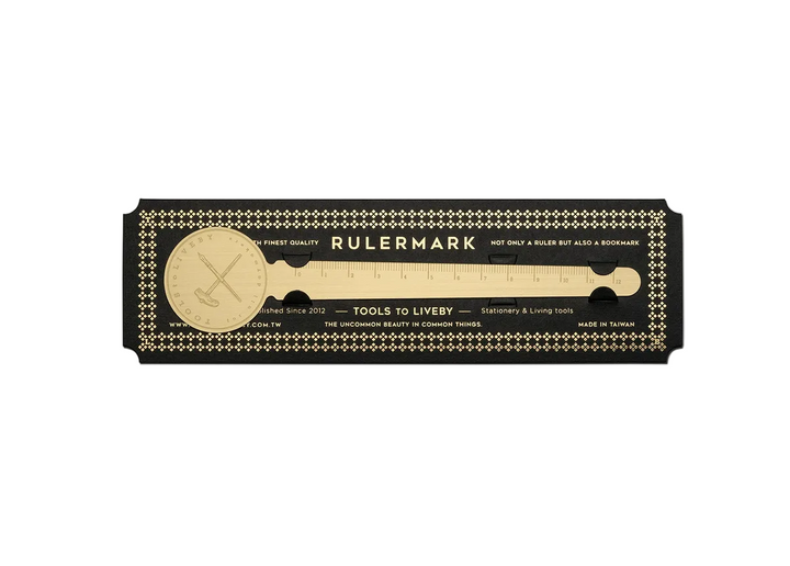 A clean, direct top-down product shot of the TOOLS to LIVEBY Brass Rulermark securely held within its premium black cardstock carrier, featuring vintage gold foil typography and a repeating diamond border against a stark white background.