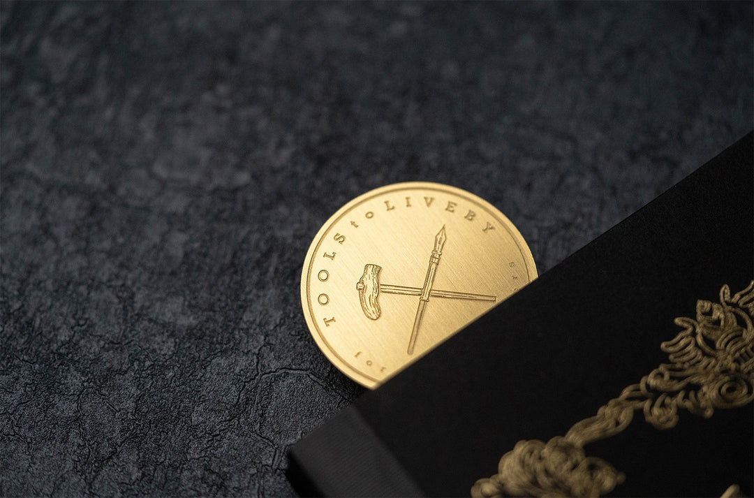 A macro detail shot of the TOOLS to LIVEBY Brass Rulermark peeking out of its black packaging on a dark slate background, showcasing the precise engraving of the brand's iconic crossed fountain pen and hammer logo.
