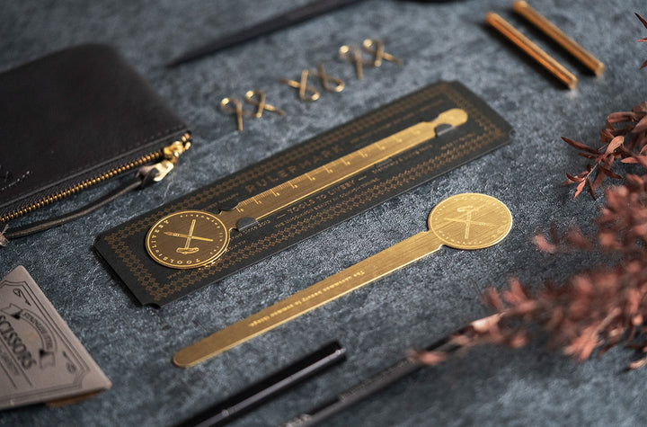 A beautifully styled dark desk aesthetic featuring the TOOLS to LIVEBY Brass Rulermark on its premium black and gold-foil packaging, surrounded by a black leather pouch, black scissors, brass paper clips, and dried botanicals.
