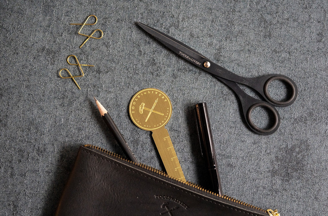 A top-down lifestyle photograph showing the TOOLS to LIVEBY Brass Rulermark tucked elegantly into a black leather zippered pouch alongside premium black writing instruments and matte black scissors on a dark textured surface.