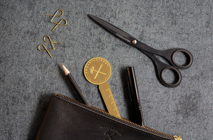 A top-down lifestyle photograph showing the TOOLS to LIVEBY Brass Rulermark tucked elegantly into a black leather zippered pouch alongside premium black writing instruments and matte black scissors on a dark textured surface.