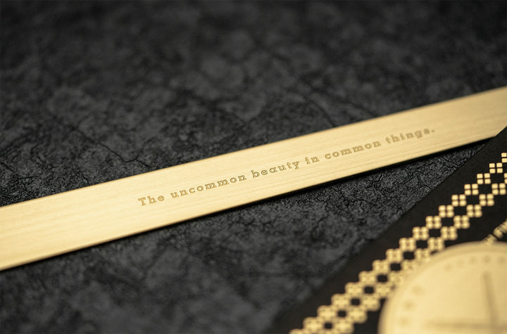 Extreme close-up of the solid brass TOOLS to LIVEBY Rulermark resting on a dark textured surface, highlighting the finely engraved motto "The uncommon beauty in common things." alongside its premium black packaging.