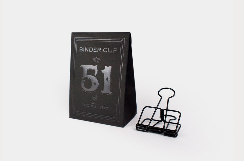 A single large 51mm TOOLS to LIVEBY matte black skeleton binder clip stands on a white surface next to its custom, black paper box, which features a prominent silver foil-stamped number '51' and brand typography.