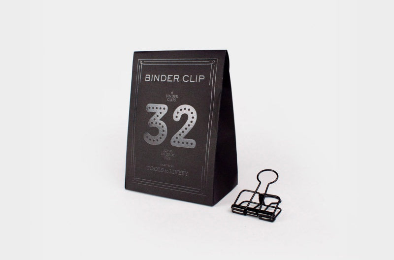 A single medium 32mm TOOLS to LIVEBY matte black skeleton binder clip stands on a white surface next to its custom, black paper box, which features a distinct dotted silver foil-stamped number '32' and brand typography.
