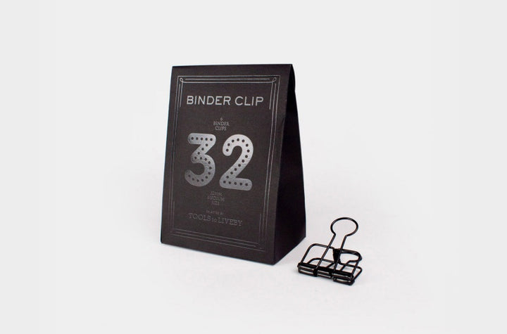 A single medium 32mm TOOLS to LIVEBY matte black skeleton binder clip stands on a white surface next to its custom, black paper box, which features a distinct dotted silver foil-stamped number '32' and brand typography.