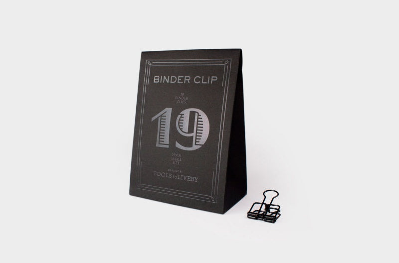 A single small 19mm TOOLS to LIVEBY matte black skeleton binder clip stands on a white surface next to its custom, black paper box, which features a distinct striped silver foil-stamped number '19' and brand typography.