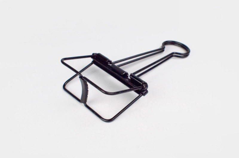 A detailed close-up shot of a single TOOLS to LIVEBY matte black skeleton binder clip lying flat against a plain white background, angled slightly to show the bottom base construction.