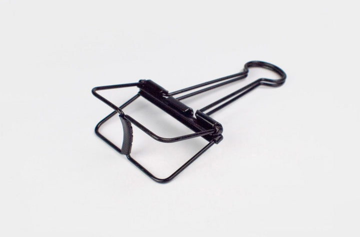 A detailed close-up shot of a single TOOLS to LIVEBY matte black skeleton binder clip lying flat against a plain white background, angled slightly to show the bottom base construction.