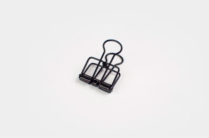 A detailed front-facing close-up shot of a single, smaller TOOLS to LIVEBY matte black skeleton binder clip standing on its base against a plain white background, displaying its robust wireframe structure.