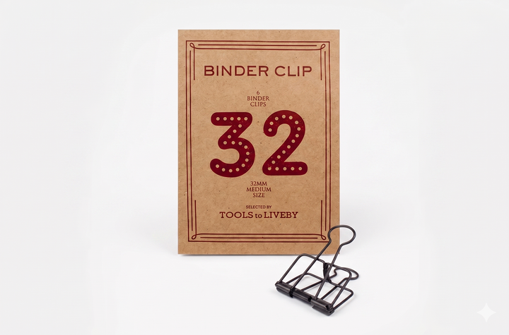 A medium 32mm TOOLS to LIVEBY Skeleton Binder Clip in bronze positioned perfectly in front of its custom foil-stamped 32mm kraft paper packaging.