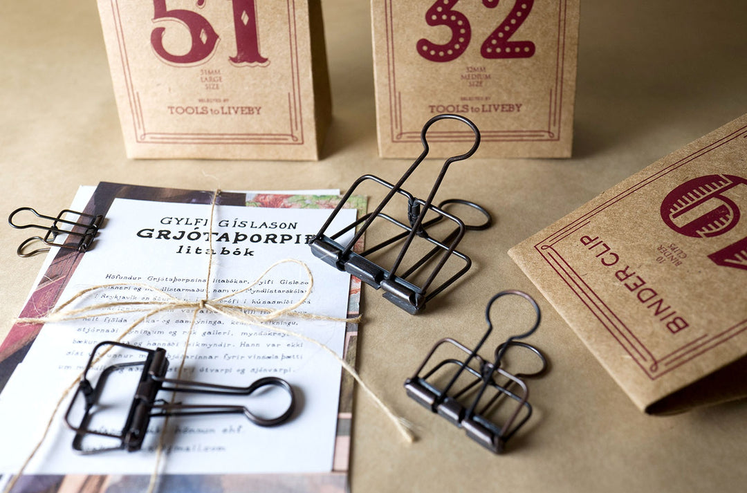 An analog stationery flatlay showing bronze TOOLS to LIVEBY Skeleton Binder Clips holding a twine-wrapped document, surrounded by their vintage kraft paper packaging.