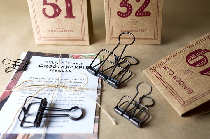 An analog stationery flatlay showing bronze TOOLS to LIVEBY Skeleton Binder Clips holding a twine-wrapped document, surrounded by their vintage kraft paper packaging.