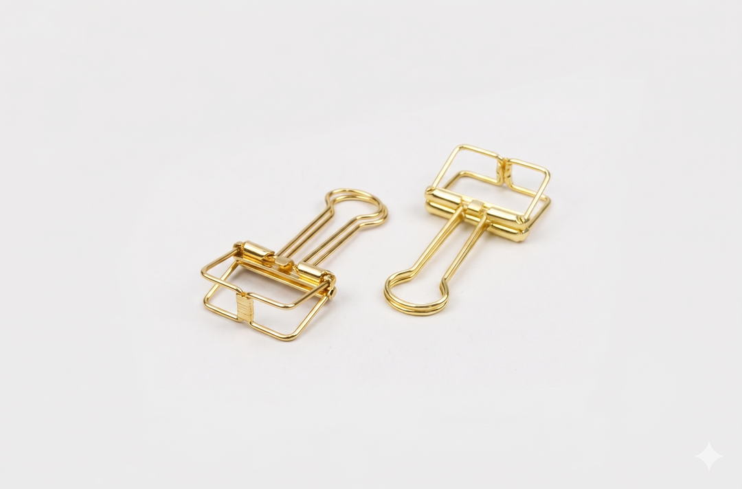 A top-down studio shot of two gold TOOLS to LIVEBY skeleton binder clips lying flat on a white surface, clearly displaying the sleek wireframe design and durable spring steel construction.
