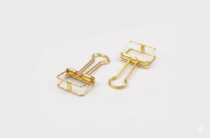 A top-down studio shot of two gold TOOLS to LIVEBY skeleton binder clips lying flat on a white surface, clearly displaying the sleek wireframe design and durable spring steel construction.