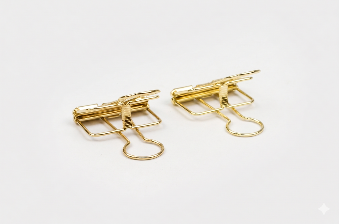 A close-up side angle of two gold TOOLS to LIVEBY skeleton binder clips resting on a white background, highlighting the luxurious gold finish and architectural tension mechanism.