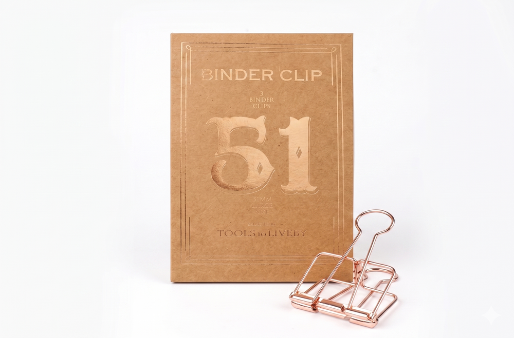 A large 51mm TOOLS to LIVEBY Skeleton Binder Clip in a luxurious Rose Gold finish, displayed alongside its sophisticated, vintage-inspired foil-stamped kraft paper box which prominently features the number '51'.