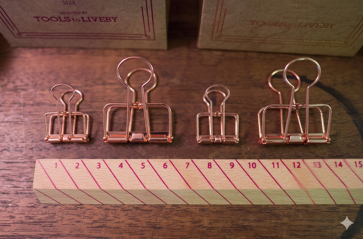 A lineup of four Rose Gold TOOLS to LIVEBY Skeleton Binder Clips arranged above a stylish striped wooden ruler on a warm wood-grain desk surface, showcasing the scale of small and large sizes in a productive workspace setting.