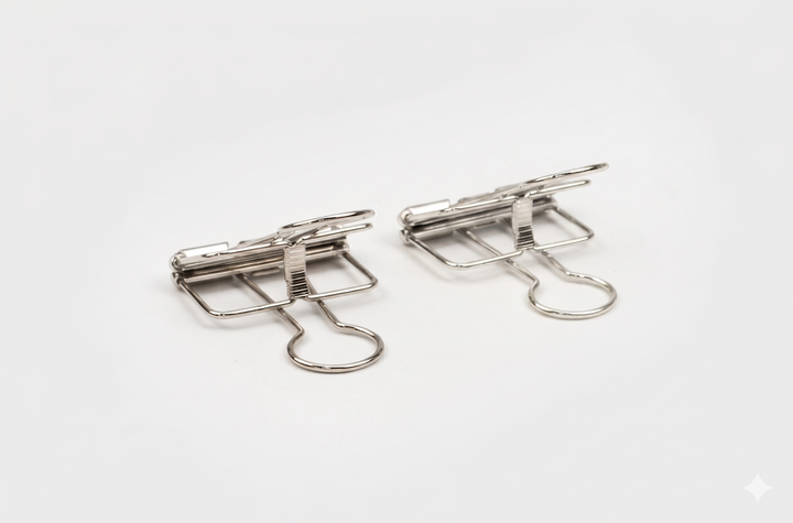 A close-up side angle of two silver TOOLS to LIVEBY skeleton binder clips resting on a white background, highlighting the high-polish chrome finish and architectural tension mechanism.
