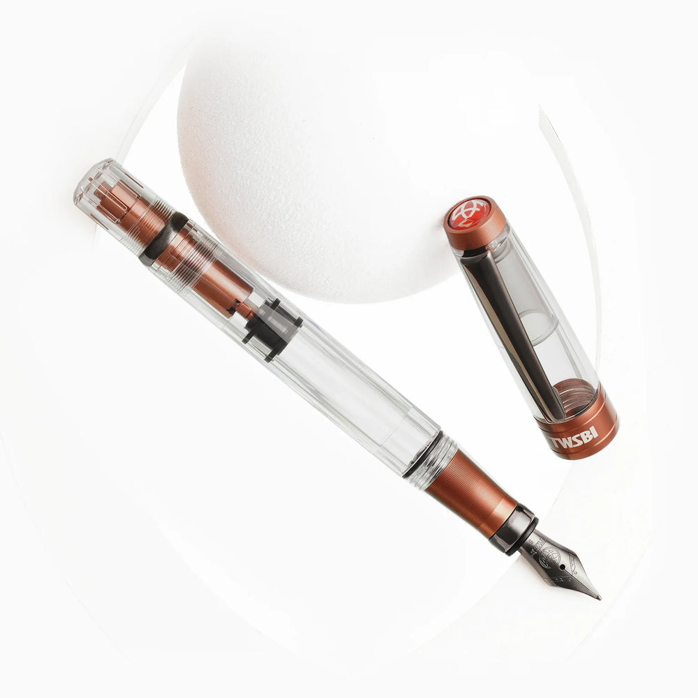 Artistic display of the TWSBI Diamond 580ALR Affogato fountain pen and cap resting against white geometric shapes, emphasizing the clear faceted barrel, black titanium clip, and matte coffee-colored trim.