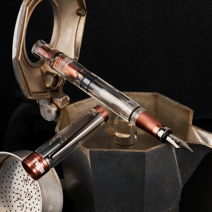 Dramatic product shot of the uncapped TWSBI Diamond 580ALR Affogato fountain pen resting on a vintage metal moka pot against a dark background, showcasing the stealthy black titanium nib and ribbed brown aluminum grip.
