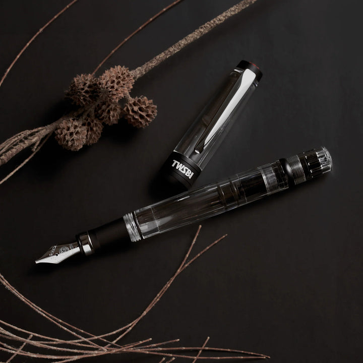 Dark and moody flat lay of the TWSBI Diamond 580ALR Black fountain pen next to its cap, arranged with dried botanicals and branches, emphasizing the stealthy matte black aluminum trim and clear faceted barrel.