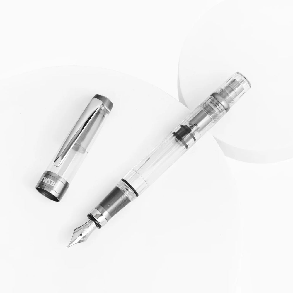 Artistic flat lay of the uncapped TWSBI Diamond 580ALR Nickel Gray fountain pen and its cap arranged on white geometric display blocks. The bright white background emphasizes the pen's clear faceted resin barrel and the sleek, modern contrast of the matte nickel-gray anodized aluminum accents.