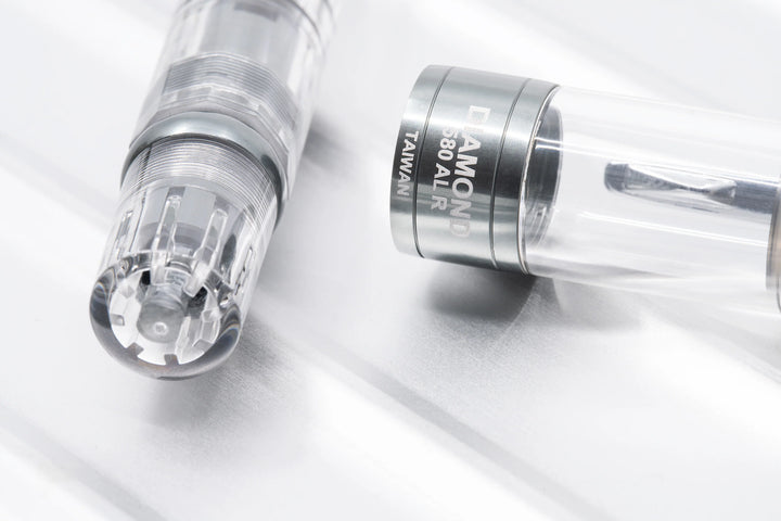 Macro close-up of the TWSBI Diamond 580ALR Nickel Gray fountain pen resting on a ridged metallic surface, sharply focusing on the clear blind cap revealing the internal piston screw, alongside the detached cap showing the engraved 'DIAMOND 580 AL R TAIWAN' text on the matte nickel-gray cap band.