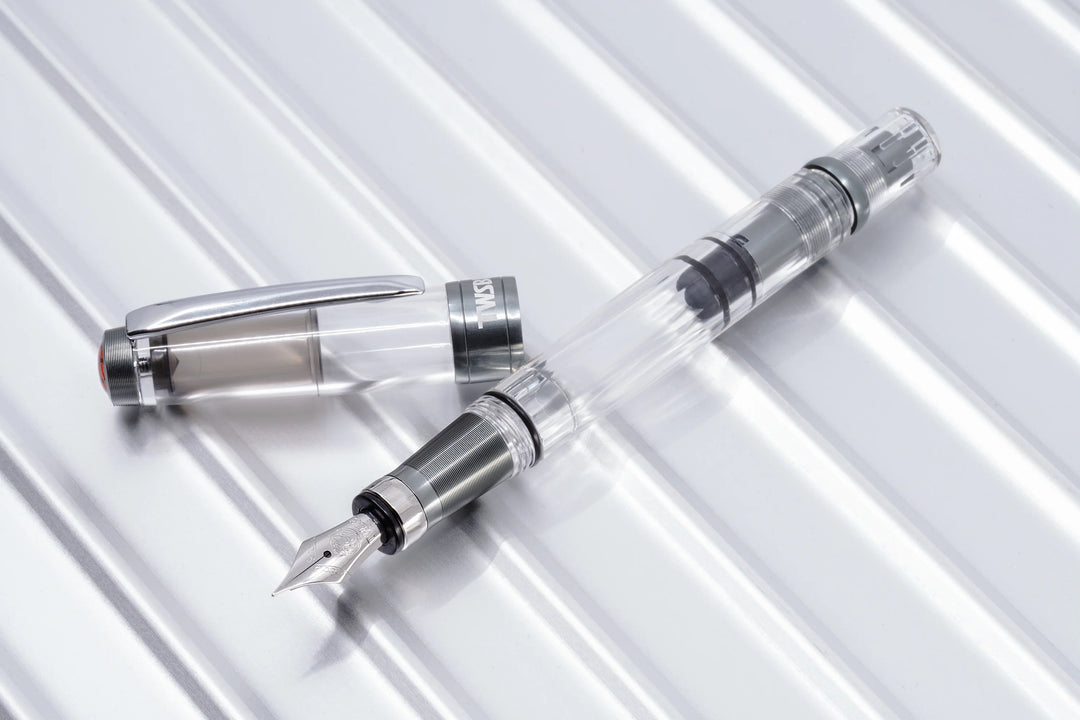 Diagonal top-down view of the uncapped TWSBI Diamond 580ALR Nickel Gray fountain pen and its cap resting on a polished, ridged metallic background. The image highlights the clear faceted demonstrator barrel, silver stainless steel Jowo nib, and the industrial matte nickel-gray ribbed aluminum grip and trim.