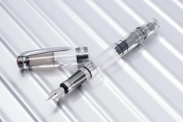 Diagonal top-down view of the uncapped TWSBI Diamond 580ALR Nickel Gray fountain pen and its cap resting on a polished, ridged metallic background. The image highlights the clear faceted demonstrator barrel, silver stainless steel Jowo nib, and the industrial matte nickel-gray ribbed aluminum grip and trim.
